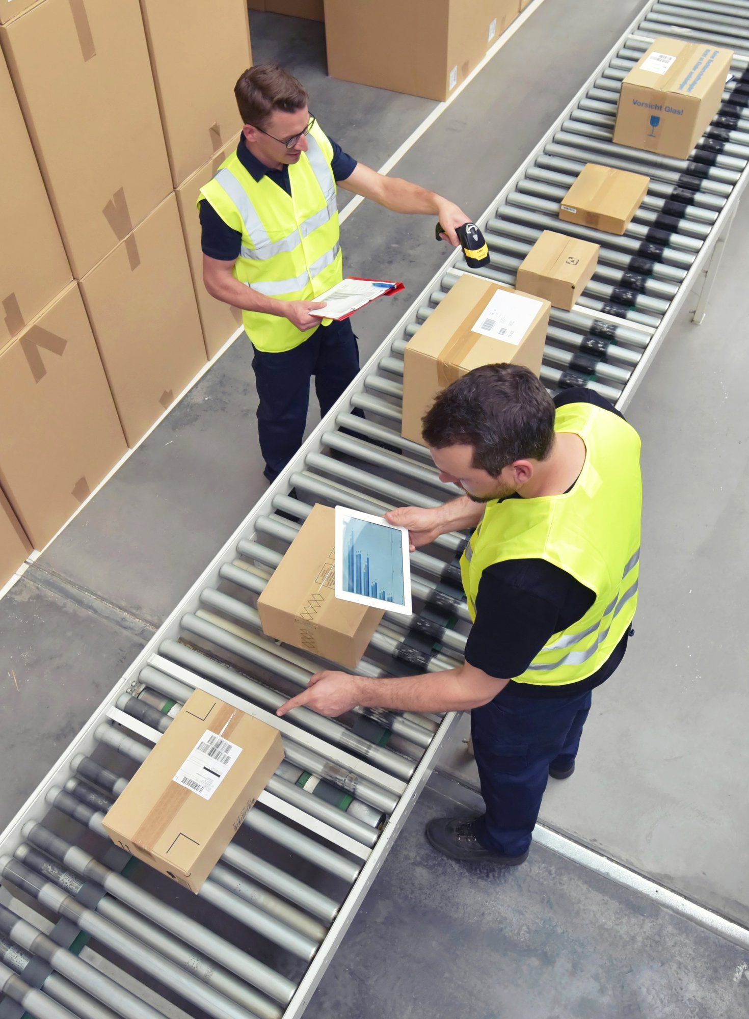Two warehouse workers in high-visibility vests manage packages on a conveyor belt—one scans a box with a handheld barcode scanner while holding a clipboard, and the other uses a laptop to inspect or label packages. Stacks of cardboard boxes are visible in the background.