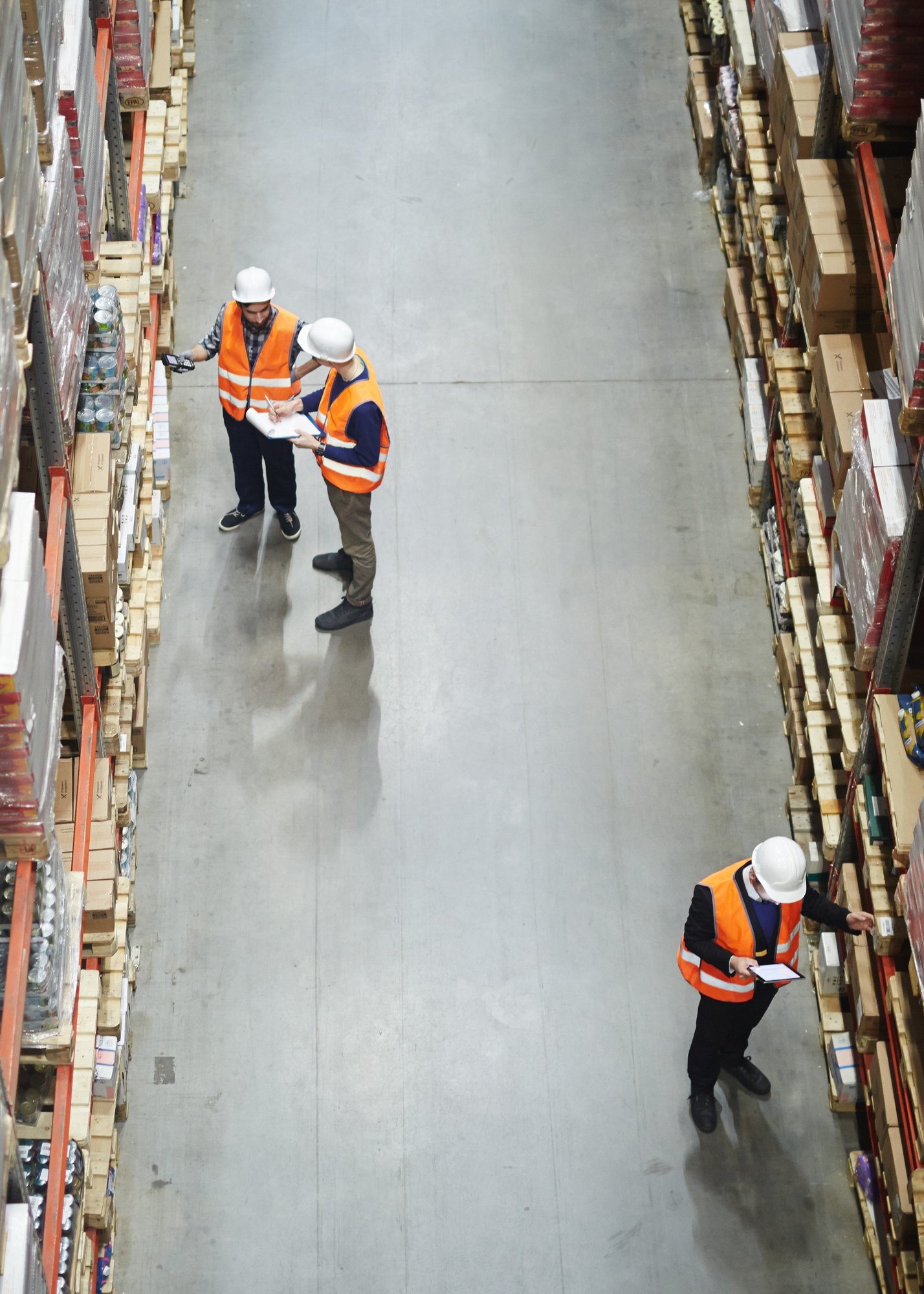 Overhead view of a warehouse aisle with four workers wearing white hard hats and orange safety vests, inspecting inventory on tall, neatly organized shelves filled with boxes and packages.